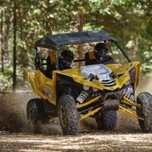 A yellow off-road vehicle with two occupants drives on a dirt path surrounded by trees.
