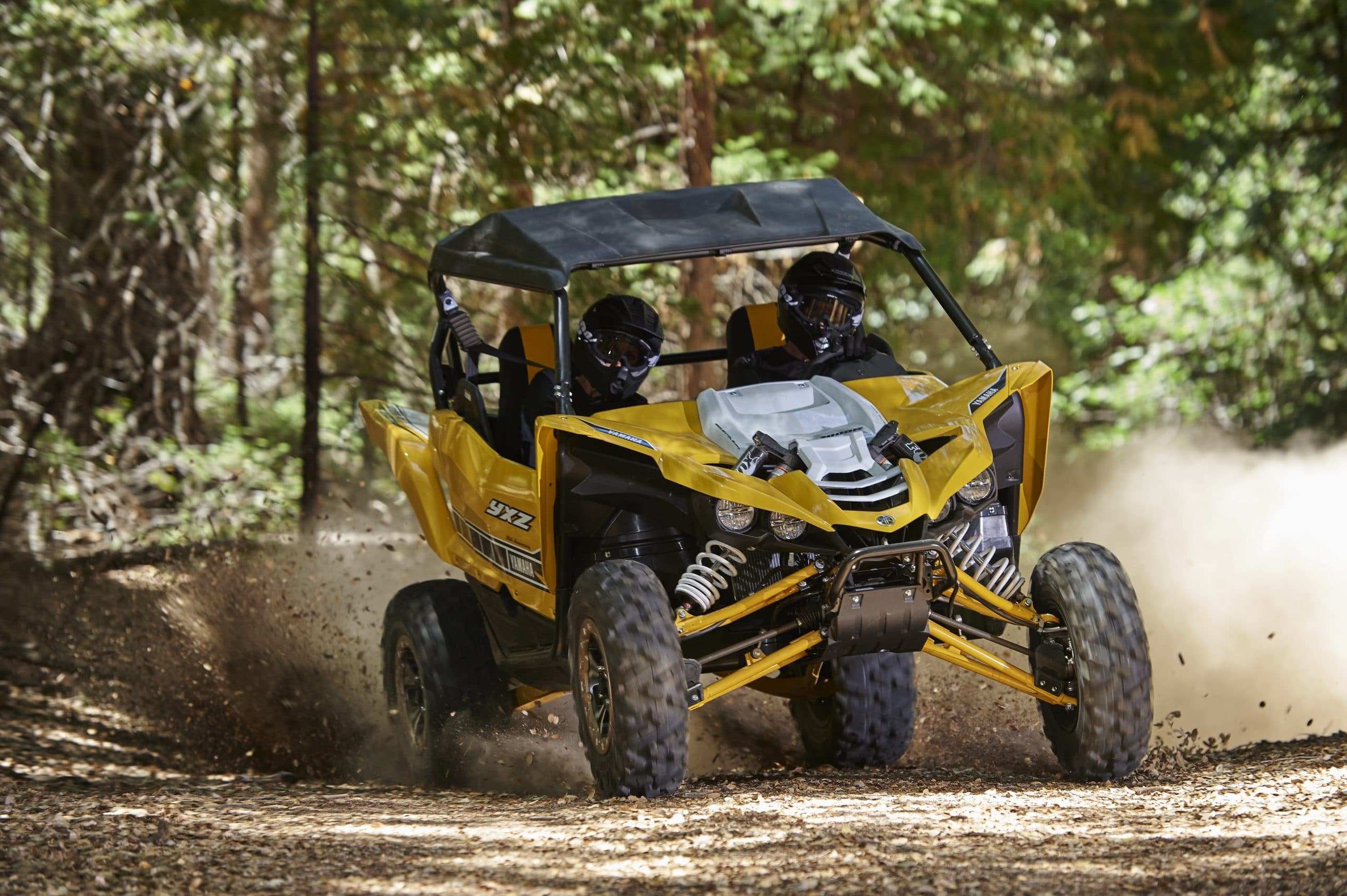 First Service: ATV (under 100cc) 3 A yellow off-road vehicle with two occupants drives on a dirt path surrounded by trees.