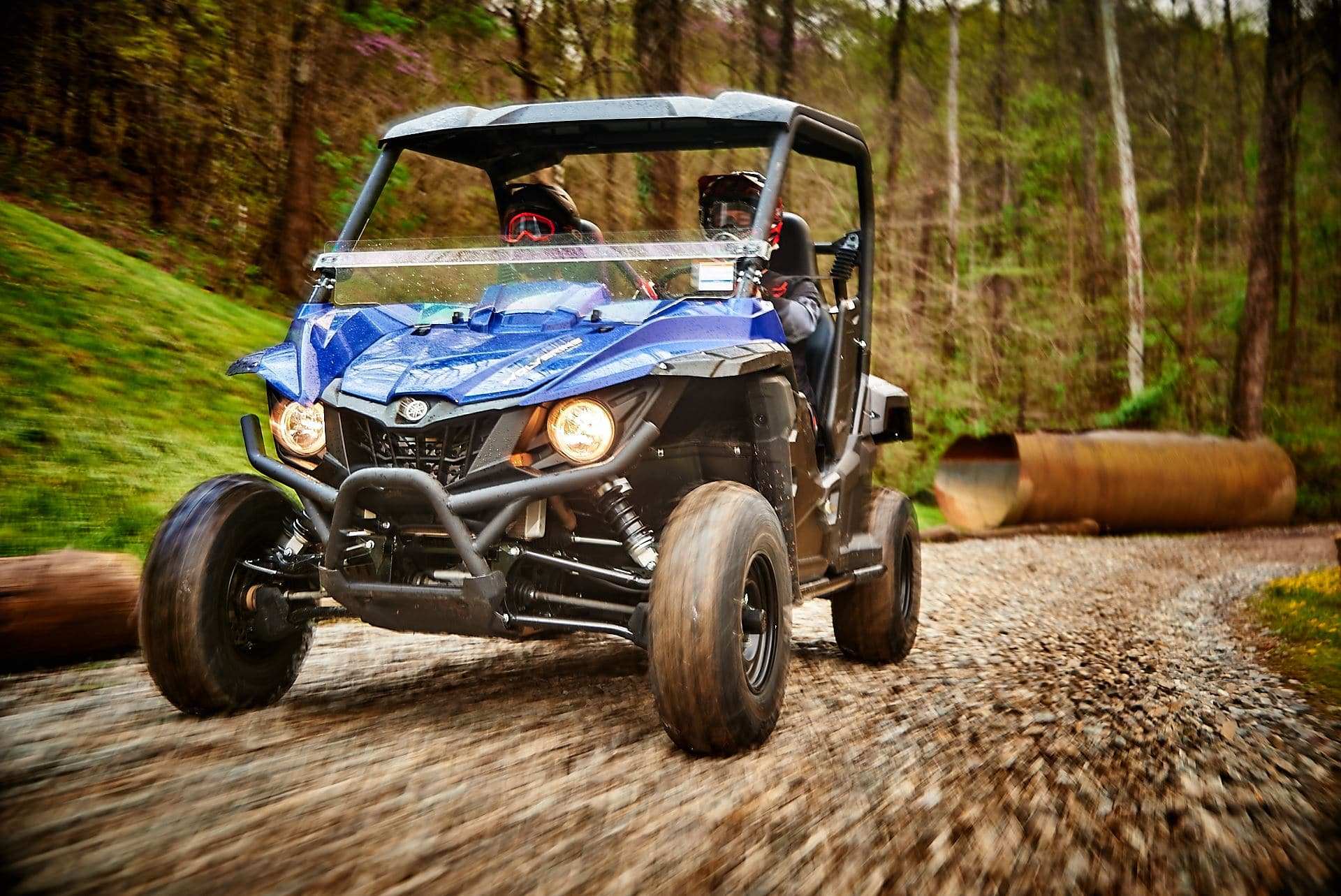 First Service: ATV (under 100cc) 5 A blue off-road vehicle with two passengers drives on a rocky path in a wooded area.