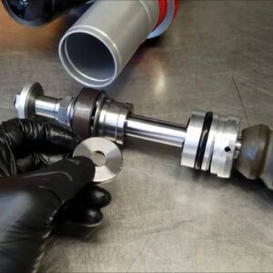 A gloved hand holds a metal ring beside a disassembled, serviceable shock absorber on a metal surface in Moorebank, Liverpool, ready for motorcycle lowering with the Lowering product.