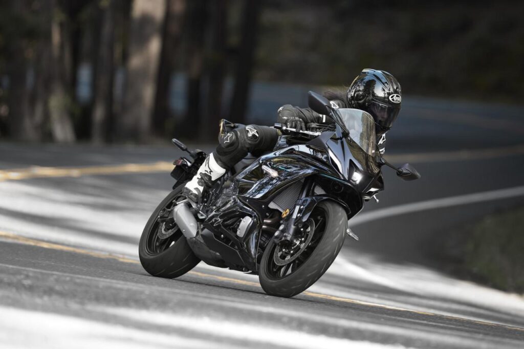 A person in full riding gear leans into a curve on a sleek, black motorcycle on a paved, winding road.