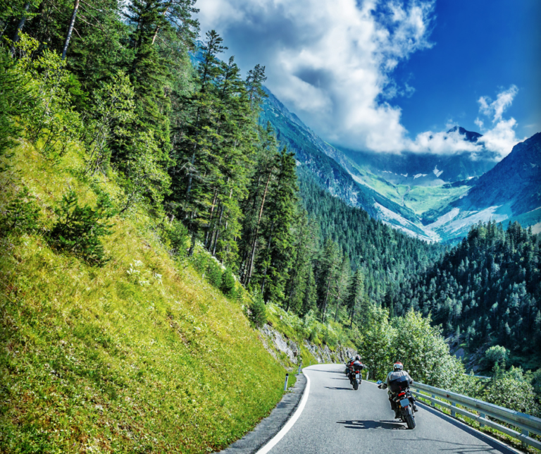 Two motorcyclists ride along a winding mountain road, flanked by lush green forests and under a sky with scattered clouds.