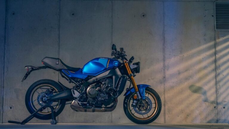 A blue motorcycle on a stand is parked indoors against a concrete wall with subtle lighting.