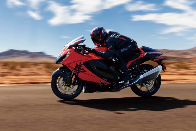 Motorcyclist in red and black gear riding a red sportbike on an open road with desert landscape in the background.