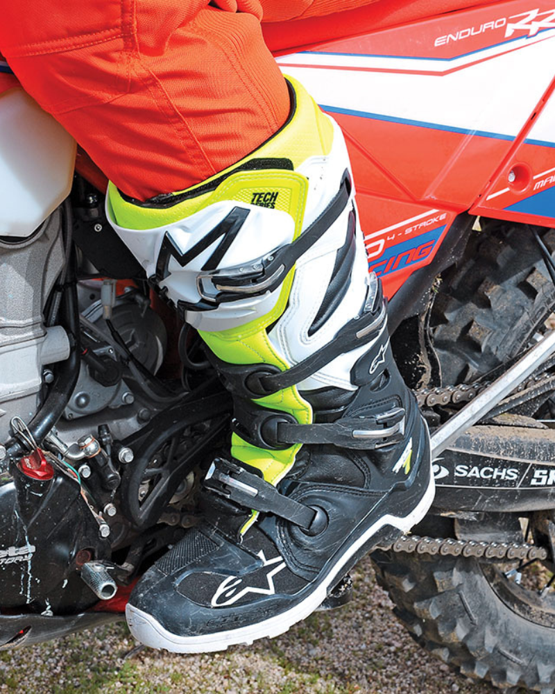 Close-up of a person wearing black, white, and yellow motocross boots, sitting on a motorcycle with red and white bodywork.