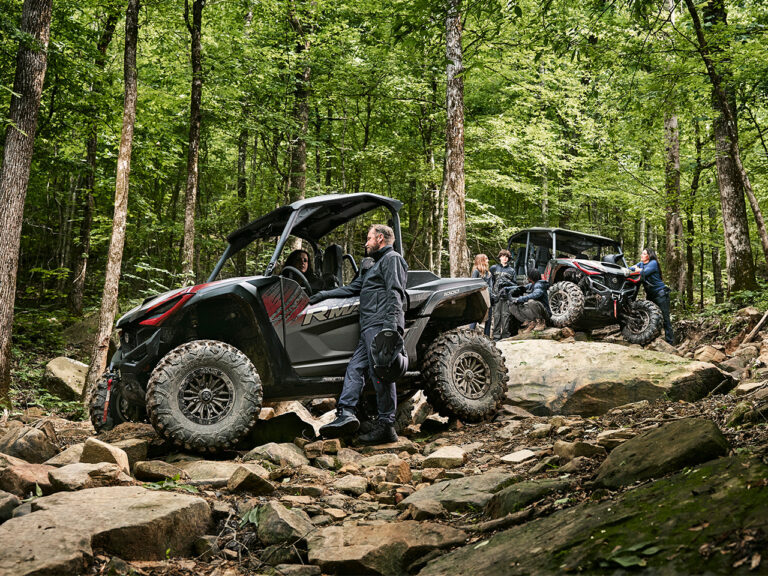 Two off-road vehicles, including a Yamaha Wolverine RMAX2 1000, navigate a rocky trail in a dense forest. Drivers and passengers dressed in outdoor gear are clearly visible as they tackle the rugged terrain.