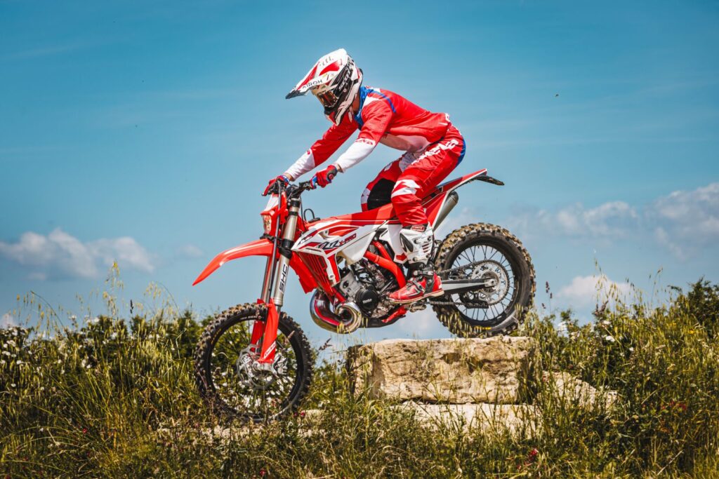 A person clad in red and white gear commands a red dirt bike, possibly a Beta motorcycle, soaring over a mound of earth with grassy accents and a backdrop of cloudy blue skies.