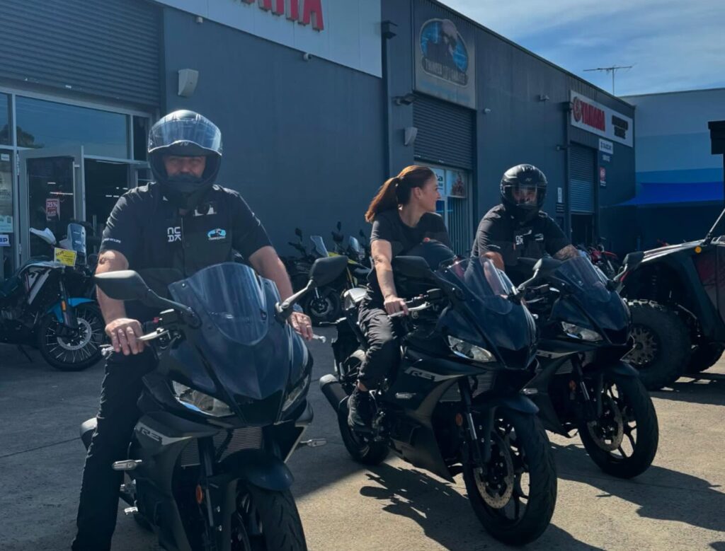 Three people wearing helmets sit on motorcycles outside a bustling motorcycle showroom in Sydney on a sunny day.