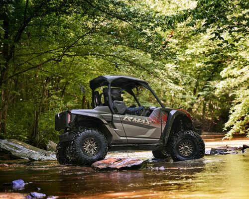 A Yamaha Wolverine RMAX2 1000 is parked on rocky terrain, surrounded by a lush forest with green trees, embodying the spirit of off-road adventure.