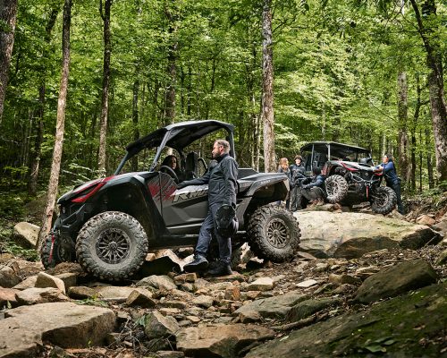 Two off-road vehicles, including a Yamaha Wolverine RMAX2 1000, navigate a rocky trail in a dense forest. Drivers and passengers dressed in outdoor gear are clearly visible as they tackle the rugged terrain.