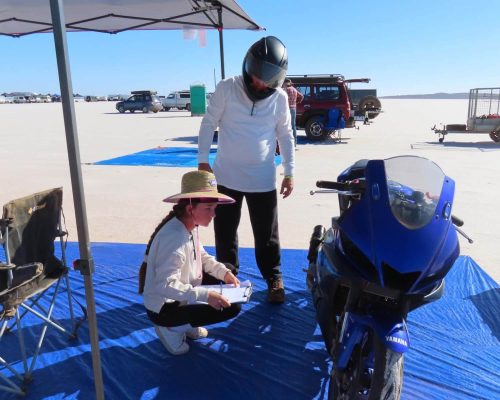 A person wearing a helmet stands next to a blue Yamaha YZF-R3 under a canopy. Another individual kneels with a clipboard, surrounded by vehicles and trailers scattered across the flat, open terrain.