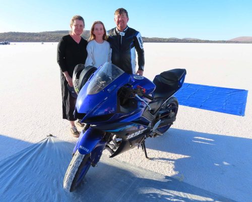 Three people stand on a vast salt flat behind a sleek blue Yamaha YZF-R3 motorcycle, the sky stretching clear and blue above them.